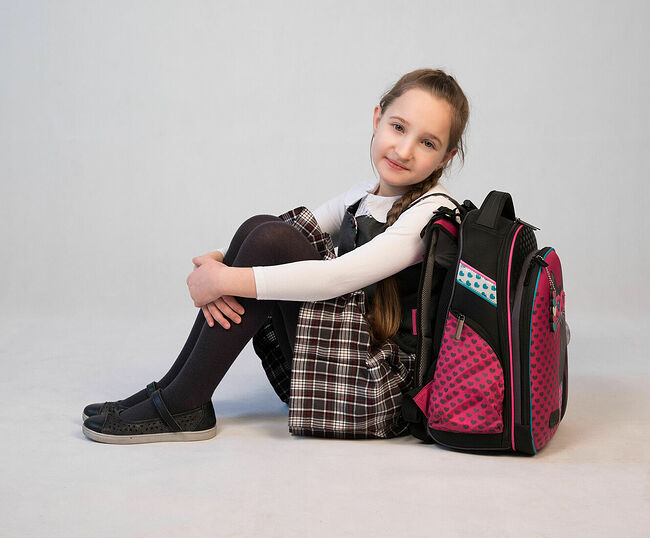 Schoolgirl sitting hugging her knees with a backpack on a light background